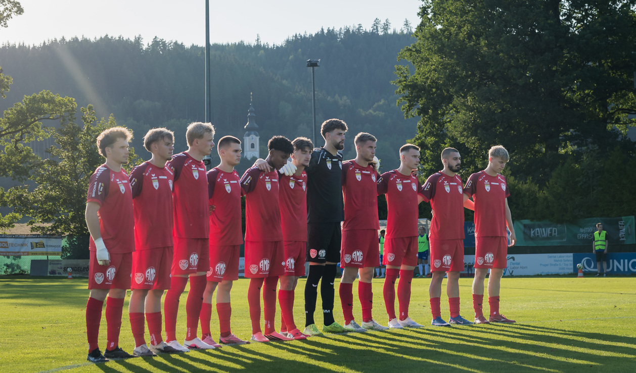 Fußballmannschaft in roten Trikots steht Spalier auf Rasen vor Bäumen und Kirche