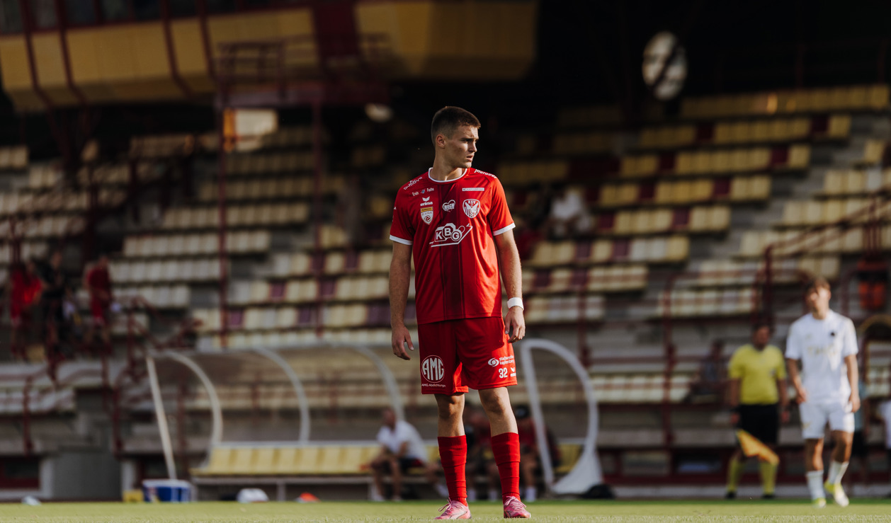 Fußballspieler in rotem Dress steht auf Rasen vor Tribüne in Kärnten